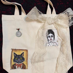 May include: Cream-colored tote bag with a cream-colored bow and lace trim. The bag features a small clock pin, a portrait of a black cat in a suit, and a portrait of a woman. The bag has cream-colored handles.