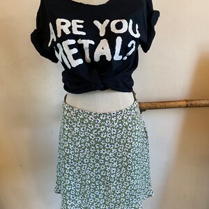 May include: A black t-shirt with the text "Are You Metal?" in white lettering. The shirt is tied at the waist and worn with a green skirt with a white floral pattern.