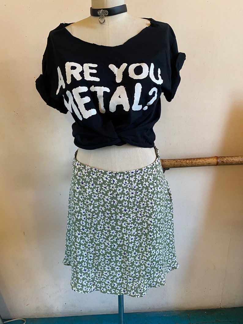 May include: A black t-shirt with the text "Are You Metal?" in white lettering. The shirt is tied at the waist and worn with a green skirt with a white floral pattern.
