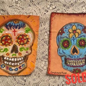May include: Two painted sugar skull art pieces on a peach background. The skull on the left is white with orange, yellow, and green details. The skull on the right is blue with orange, yellow, and green details. Both skulls have a cross on their foreheads. The text "Cashmere Funk" is written on both pieces.