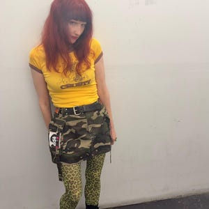 May include: A yellow short-sleeve ringer t-shirt with brown trim and the text CRUSTY printed on the front, a camouflage print mini skirt with a black belt, green and black leopard print leggings, and black mid-calf boots with silver buckles.