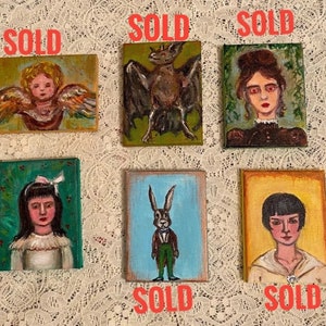 May include: Six small paintings on canvas, each with a different subject. The paintings are framed in wood and have a white background. The subjects include a bat, a rabbit, an angel, and three portraits of people. Each painting has the word "SOLD" written in red letters below it.
