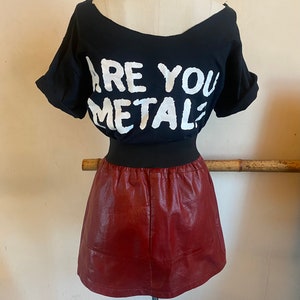 May include: A black t-shirt with the text "ARE YOU METAL?" printed in white. The t-shirt is paired with a burgundy faux leather mini skirt.