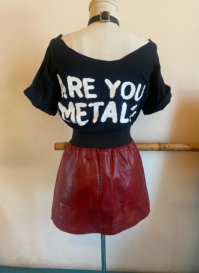 May include: A black t-shirt with the text "ARE YOU METAL?" printed in white. The t-shirt is paired with a burgundy faux leather mini skirt.