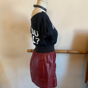 May include: A maroon faux leather mini skirt with a black elastic waistband and a button closure. The skirt is on a mannequin wearing a black t-shirt with the text "Do you feel?" in white.