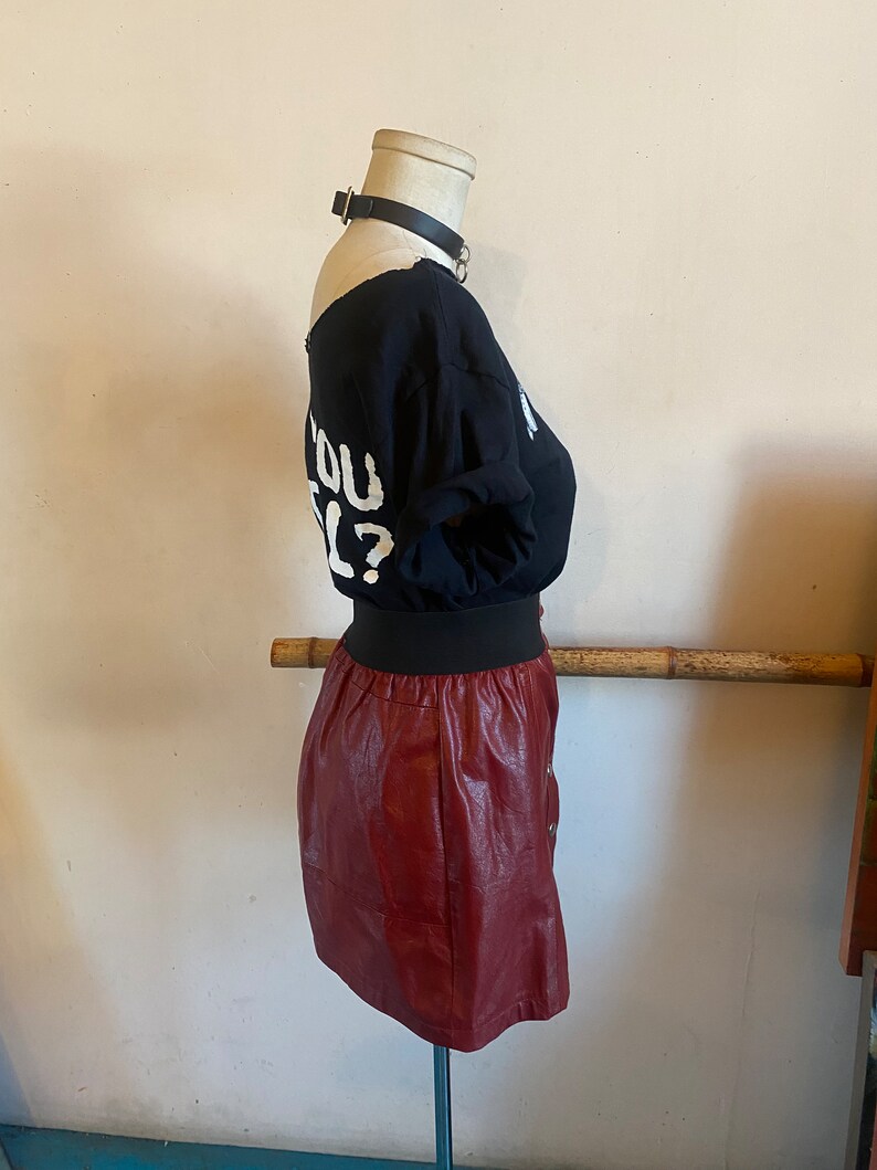 May include: A maroon faux leather mini skirt with a black elastic waistband and a button closure. The skirt is on a mannequin wearing a black t-shirt with the text "Do you feel?" in white.