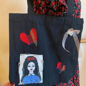 May include: Black canvas tote bag with a red and black plaid heart, a painted portrait of a woman with a red bow, and a smaller red and black plaid heart. The bag has a black strap.