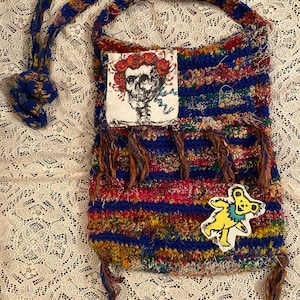 May include: A crocheted bag with a blue, red, yellow, and green striped pattern. The bag has a skull with roses and a dancing bear patch. The bag has fringe on the bottom.