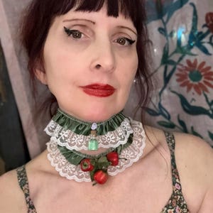 May include: A green velvet and lace choker necklace with a small glass bottle pendant and red apple accents. The choker features a layered design with white lace trim and a satin finish.
