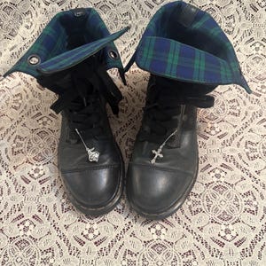 May include: A pair of black leather lace-up boots featuring a fold-over cuff with a blue and green plaid pattern. The boots have black laces and are adorned with two silver-toned metal charms, one shaped like a cross and the other a small figure.
