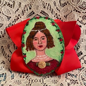 May include: A hand-painted portrait of a woman with brown hair, wearing a white headband and a red dress with a green rose brooch. The portrait is painted on a round wooden surface and is surrounded by a green border with red roses.