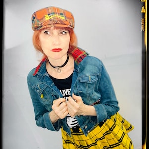 May include: A woman wearing a yellow and black plaid skirt, a denim jacket, a black shirt with the word "LIVE" on it, and a plaid newsboy cap.