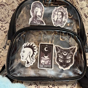 May include: A clear, black-trimmed backpack adorned with stickers. Stickers include a crying face, a portrait, a punk with a mohawk, a tarot card with the text "THE COFFEE", and a cat with multiple eyes.