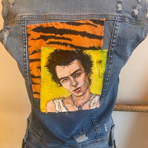 May include: A sleeveless, distressed denim jacket with a tiger stripe patch and a portrait of a person on a yellow background. The portrait is secured with safety pins. The jacket is worn over a plaid skirt.