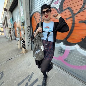 May include: A person wearing a light blue top with a graphic design, a black cardigan, and a plaid skirt. They are also wearing black tights and shoes. A gray bag with the word "MIN" is hanging from their shoulder. The background features graffiti art.