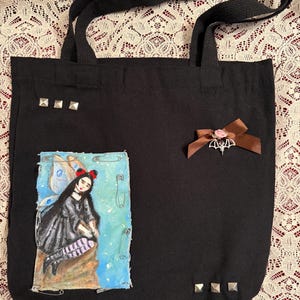 May include: Black canvas tote bag with a painted image of a fairy with blue wings and a black dress. The bag has silver studs and a brown ribbon with a bat charm.