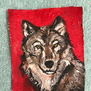 May include: A painting of a wolf's head on a red background. The wolf has a gray and brown coat with white fur around its neck. The wolf's eyes are yellow and it is looking directly at the viewer.