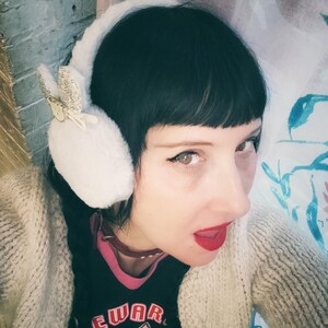 May include: White faux fur earmuffs with a decorative butterfly accent. The earmuffs are worn with a black t-shirt with the text "EWAR" and a cat graphic. A beige cardigan is also worn.