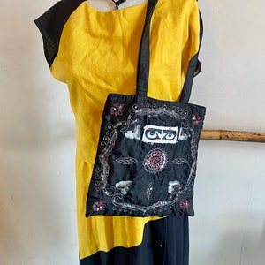May include: A mannequin wearing a yellow and black dress, a black hat, and a black beaded necklace. A black tote bag with an embroidered design featuring eyes and floral elements is hanging from the mannequin's shoulder.