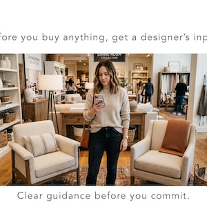 May include: A woman in a beige sweater and black jeans looks at her phone in a furniture store. Two armchairs with cream-colored upholstery and throw pillows are in the foreground. The text reads: "Before you buy anything, get a designer's input."