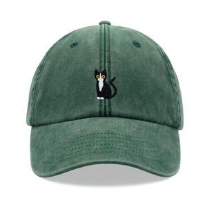 May include: A green baseball cap with an embroidered black cat with white markings and yellow eyes.