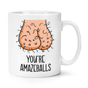 May include: White ceramic mug with a humorous illustration of a pair of testicles and the text "YOU'RE AMAZEBALLS" in black. The illustration is in a cartoon style.