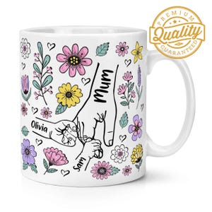 以下が含まれることがあります： 白いセラミック製のマグカップに、花柄のデザインと「Mum」、「Olivia」、「Sam」という黒い文字が書かれています。デザインは、指が絡み合った手のひら型で、家族を表しています。