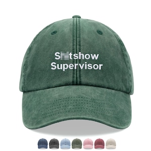 May include: A green, distressed, cotton baseball cap with the text "Sh*tshow Supervisor" embroidered in white on the front.