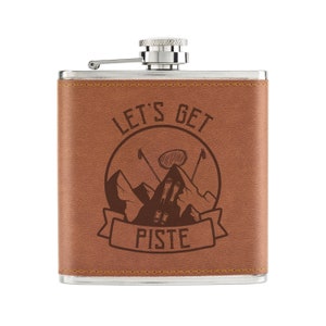 May include: A brown leather-wrapped stainless steel hip flask with a silver cap. The flask has an engraved design of mountains, ski poles, and the text "Let's Get Piste".