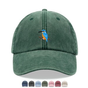 May include: A dark green baseball cap with a curved peak. The cap has an embroidered image of a blue and orange bird on a branch. Several other cap colours are shown below the main cap.
