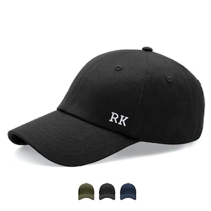May include: A black baseball cap featuring the white embroidered letters RK on the front panel. The cap has a curved brim, a structured crown with ventilation eyelets, and a button on top. Three smaller colour variations are shown below.