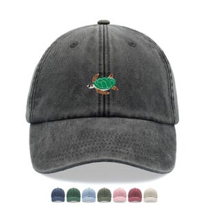 Può includere: Un cappellino da baseball grigio antracite con una piccola tartaruga marina ricamata sul davanti. La tartaruga ha un guscio verde e un corpo marrone. Il cappellino ha una visiera curva e una chiusura con cinturino in tessuto. Sotto sono mostrati cappellini colorati.