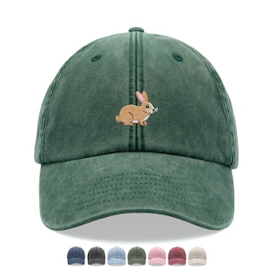 May include: A dark green baseball cap with a distressed, washed-out finish. The front panel features an embroidered design of a brown rabbit with pink-lined ears. The cap has a curved brim and a structured crown with ventilation eyelets.