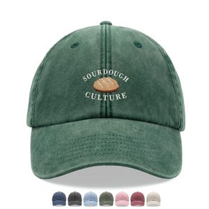 Può includere: Un cappellino da baseball verde con la scritta ricamata "SOURDOUGH CULTURE" e un disegno di pagnotta. Il cappellino ha una visiera curva e un aspetto invecchiato. Sono mostrate diverse altre opzioni di colore sotto il cappellino.