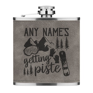 May include: A grey leather-wrapped stainless steel hip flask with a silver cap. The flask features a black design of mountains, trees, a snowboard, ski gloves, and goggles. The text "ANY NAME'S getting piste" is printed on the flask.
