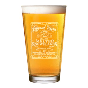 May include: A clear glass pint glass with a white design that reads "Liberal Tears" and "Quality Melted Snowflakes" with a vintage-style label. The glass is filled with a golden yellow liquid.