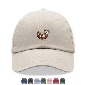 May include: A beige baseball cap with a curved brim. The cap features an embroidered brown otter design on the front. The cap is shown with a selection of six other color options at the bottom.