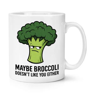 Op de afbeelding: Witte keramische mok met een cartoon broccoli illustratie en de tekst "MAYBE BROCCOLI DOESN'T LIKE YOU EITHER". De broccoli is groen met een chagrijnige uitdrukking. De mok heeft een gebogen handvat.