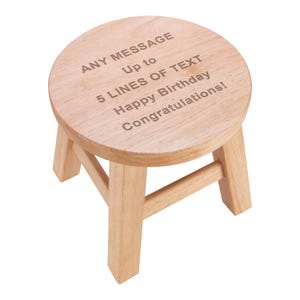 Puede incluir: Un pequeño taburete redondo de madera clara con cuatro patas. El asiento del taburete tiene grabado el texto: "ANY MESSAGE Up to 5 LINES OF TEXT Happy Birthday Congratulations!".