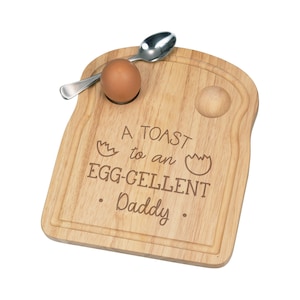 Puede incluir: Una tabla de madera para huevos y tostadas con un huevo marrón, una cuchara y el texto grabado "A TOAST to an EGG-CELLENT Daddy". La tabla tiene una hendidura circular para el huevo y una ranura alrededor del borde.