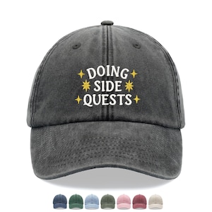 May include: A charcoal grey baseball cap with a distressed finish featuring the embroidered white text DOING SIDE QUESTS centred on the front panel, accented by six small yellow star icons arranged around the lettering in a symmetrical pattern.