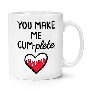 Puede incluir: Taza de cerámica blanca con el texto negro "YOU MAKE ME CUM-PLETE" sobre un corazón rojo con detalles blancos. La taza tiene un asa curva.