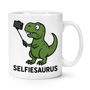 May include: White ceramic mug with a green cartoon dinosaur taking a selfie. The dinosaur has a surprised expression. The mug has the word "SELFIESAURUS" printed in black block letters.