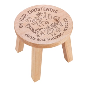 May include: A round wooden stool with three legs, featuring a laser-engraved design of a lion, giraffe, zebra, and monkey. The text reads ON YOUR CHRISTENING, AMELIA ROSE WILLIAMS, and 18.05.2025, arranged around the animal illustrations.