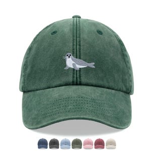 Könnte beinhalten: Grüne Baseballkappe mit einem gestickten Seehund-Design. Die Kappe hat eine einfarbige grüne Farbe mit einem gebogenen Schirm und einer sechsteiligen Konstruktion. Der Seehund ist grau und weiß. Mehrere andere Kappenfarben sind abgebildet.