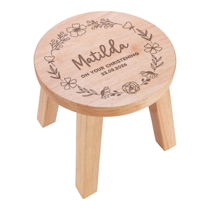 Op de afbeelding: Een kleine ronde houten kruk met drie poten. Het bovenoppervlak is voorzien van een lasergegraveerd bloemenkransontwerp rondom de naam Matilda en de tekst ON YOUR CHRISTENING 23.09.2026. Het hout heeft een lichte natuurlijke afwerking.