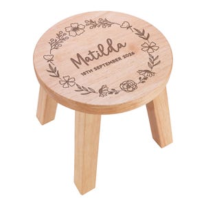 Op de afbeelding: Een ronde houten kruk met drie poten en een lasergegraveerd bloemenkransontwerp op de zitting. In het midden van de zitting staan de naam Matilda en de datum 18TH SEPTEMBER 2026 in een donkerbruin lettertype in script- en blokstijl.