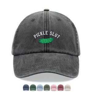 May include: A dark gray baseball cap with the embroidered text "PICKLE SLUT" above a green pickle illustration. The cap has a curved brim and a fabric strap closure. Several color options are shown below the cap.