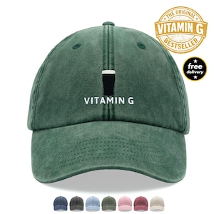 May include: Green baseball cap with white embroidered text reading "VITAMIN G". The cap displays a graphic of a dark drink in a glass. It also features a "The Original Vitamin G Bestseller" logo and a "free delivery" badge.