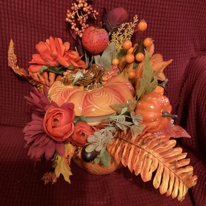 Puede incluir: Un recipiente de cerámica con forma de calabaza con un diseño de espiral está decorado con flores artificiales, hojas y bayas de otoño en tonos naranja, rojo y verde.